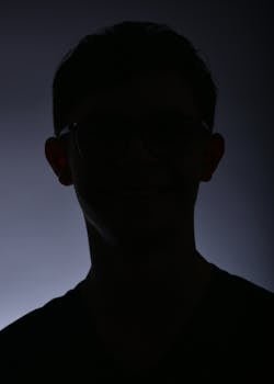 Moody silhouette portrait of a young man wearing glasses with a soft gradient background.